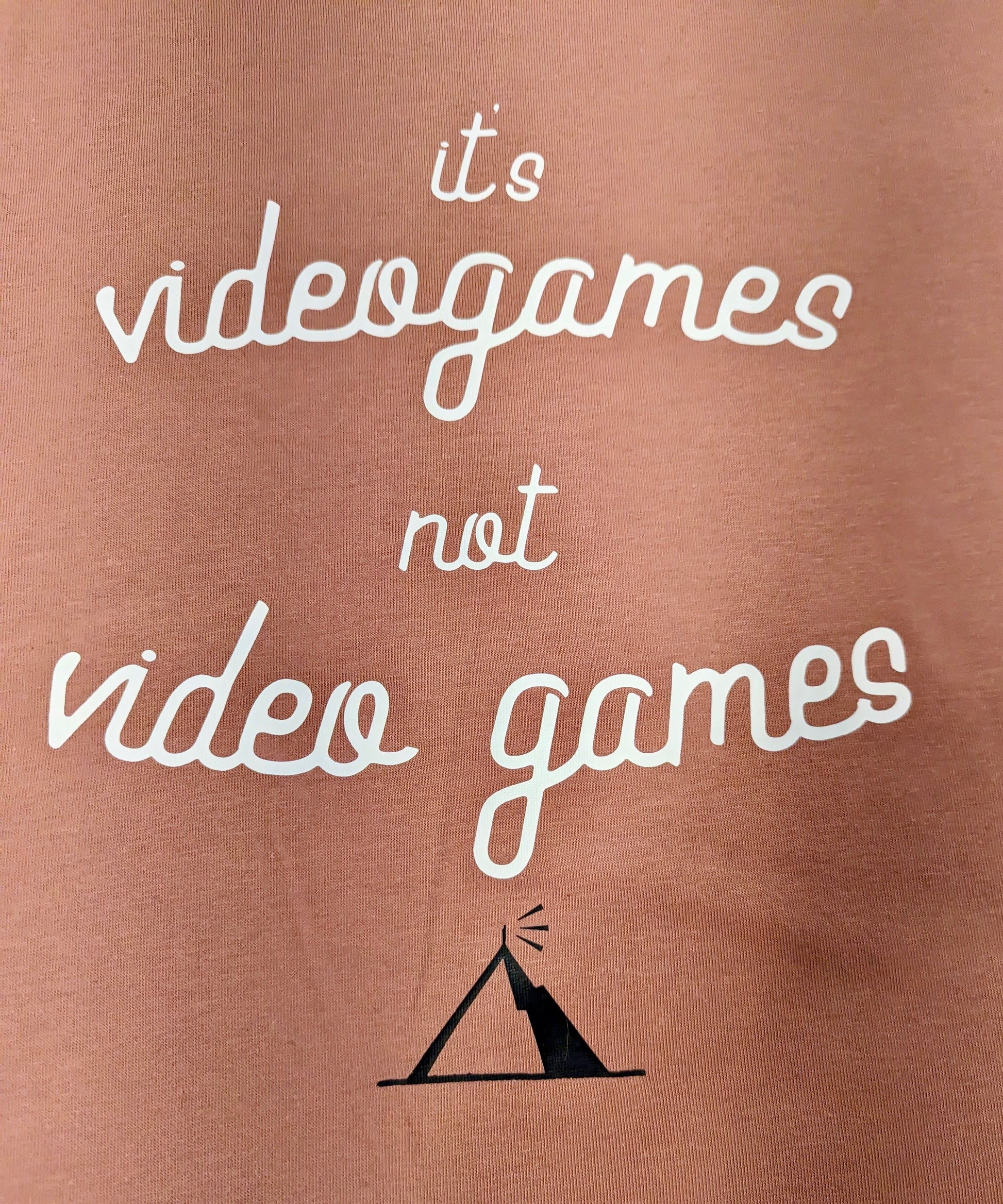 A Dusty Rose T-Shirt, with cursive writing on it in white. In lowercase letters the first line reads “it’s”, the second line “videogames” spelt as one word, the third line “not” and the fourth line “video games” using two words. In the centre below the text is the Mighty Yell logo in black which features the illustration of a mountain and a person on top yelling. There are three small lines coming out of the person’s mouth.